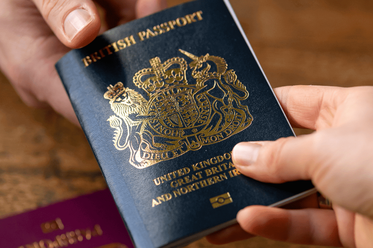 shows a photo of a dark blue with gold lettering UK passport being passed from one hand to another